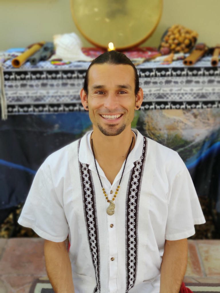 Gerardo Marin, smiling with brown hair pulled back, a goatee, white shirt, and necklace in front of an altar.